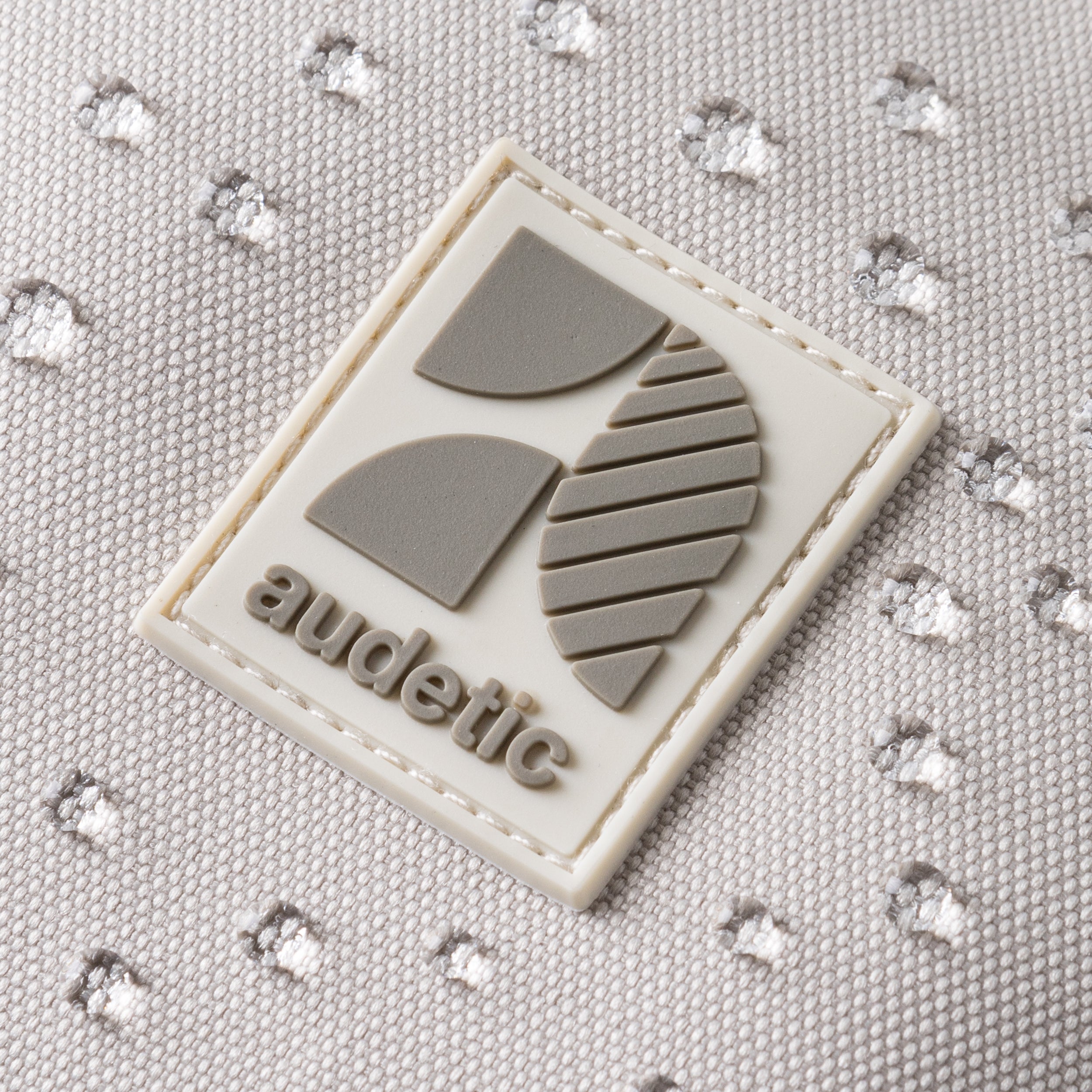 Grau-beige audetic Markenlogo auf sandfarbenem Stoff mit Wasserperlen