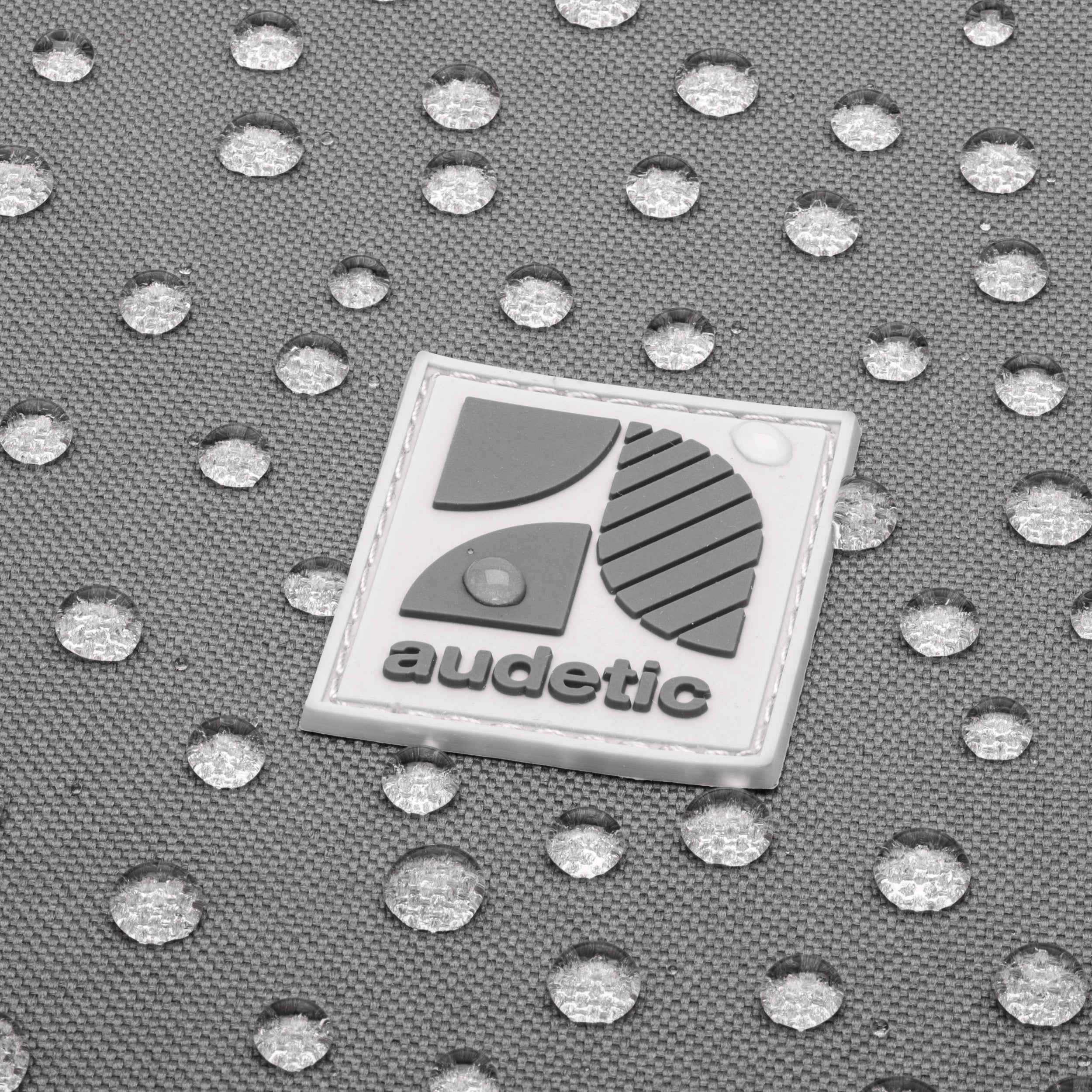 Wasserabweisendes dunkelgraues Material mit aufgenähtem weißen audetic-Logo und Wassertröpfchen