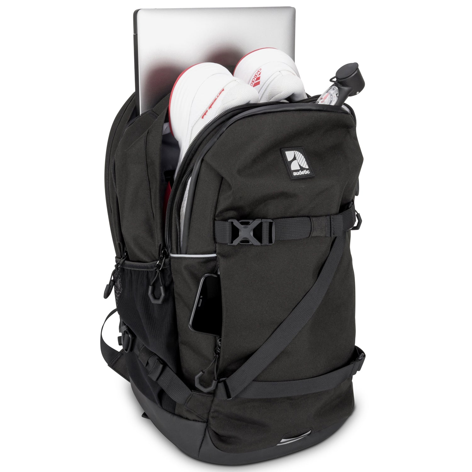 Schwarzer Schulrucksack COSMO von audetic mit geöffnetem Hauptfach, darin ein Laptop, weiße Sportschuhe und eine Kompassuhr