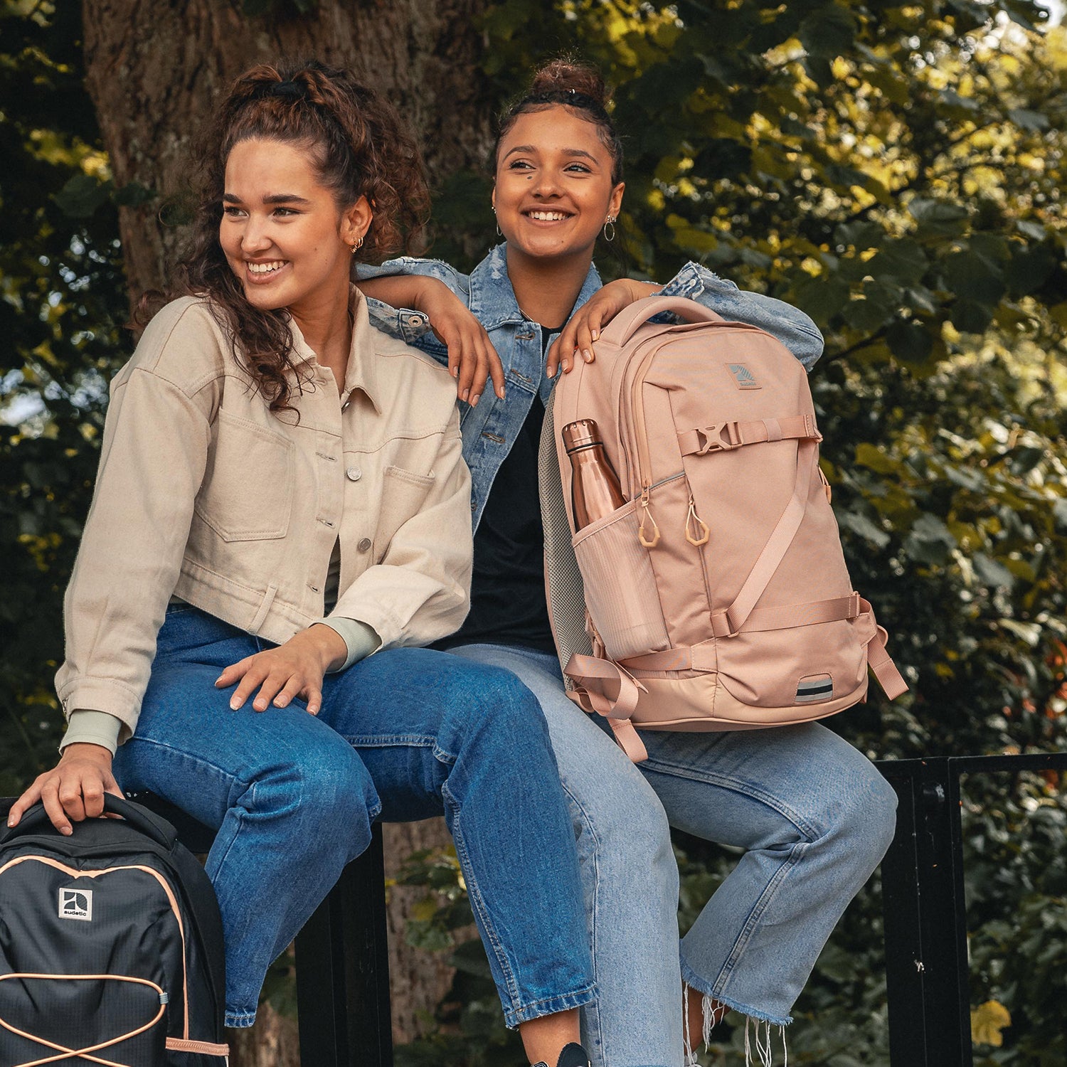 Zwei lächelnde Teenager-Mädchen sitzen draußen, eines hält einen schwarzen und das andere einen rosa Schulrucksack COSMO