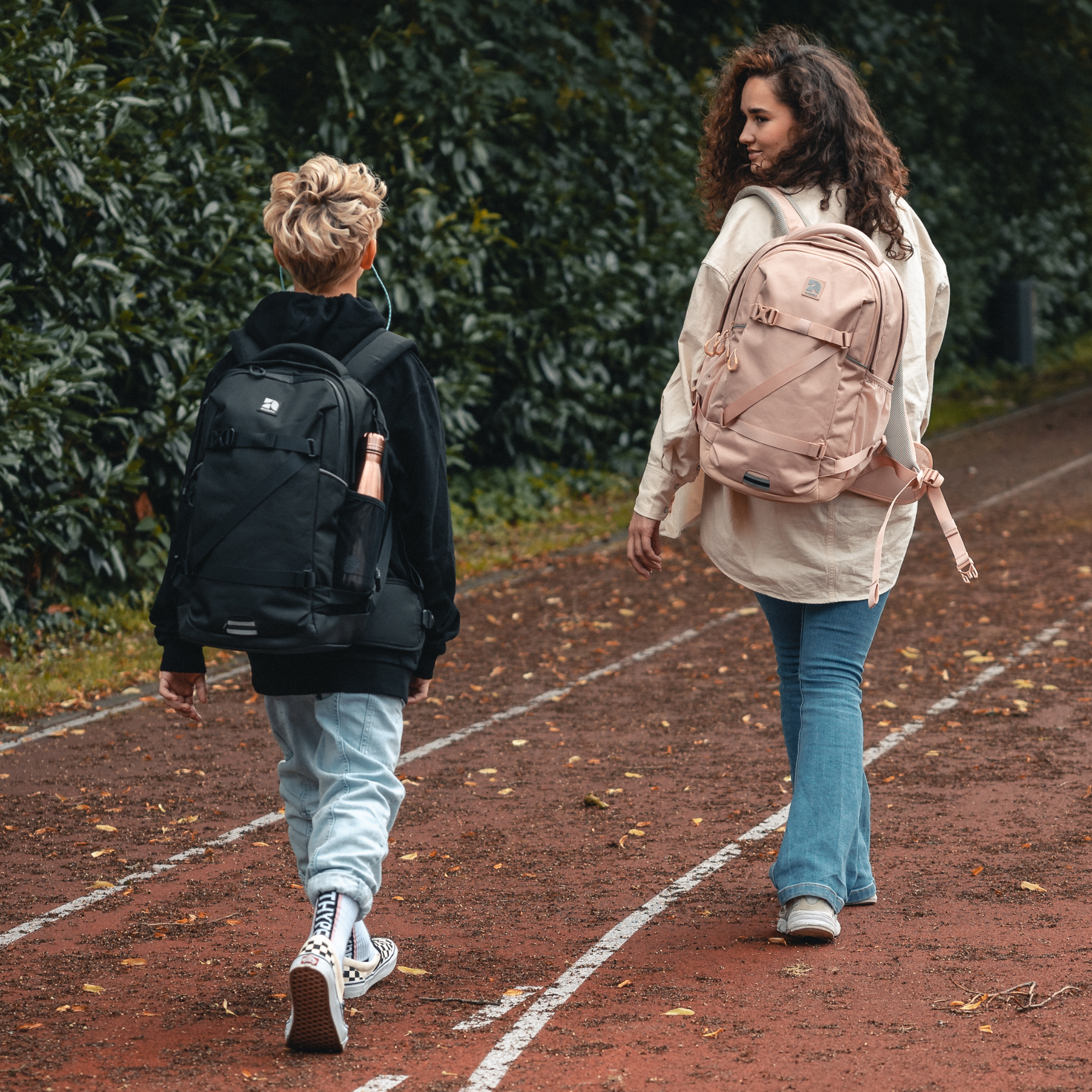 Zwei Jugendliche gehen auf einer Laufbahn, einer trägt einen schwarzen und der andere einen rosa COSMO Schulrucksack