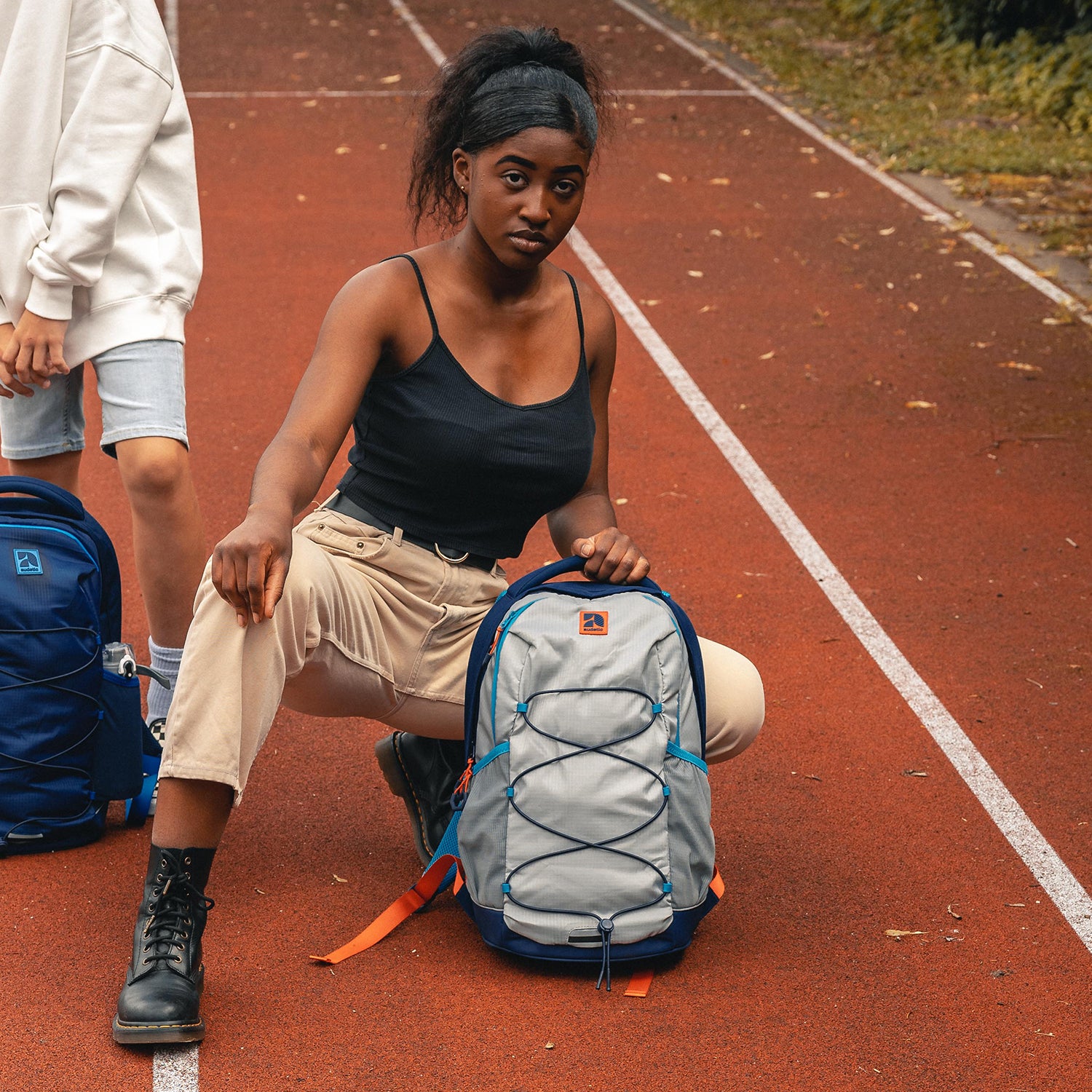 Teenager-Mädchen kniet auf roter Laufbahn und hält hellgrauen Schulrucksack AERO mit blauen und orangefarbenen Details