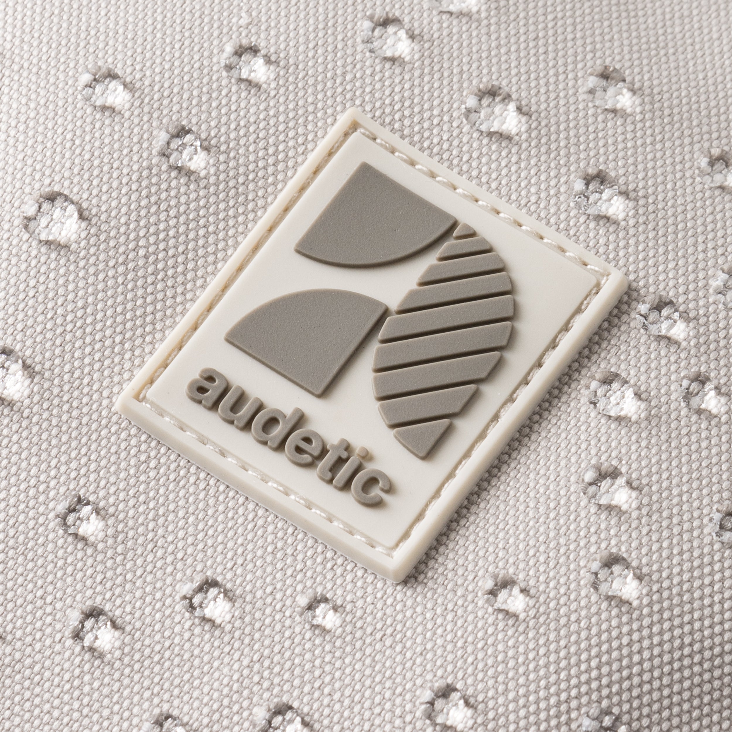 Detailaufnahme des weißen Audetic-Logos mit geometrischem Design auf hellgrauem, wasserabweisendem Schulrucksack COSMO